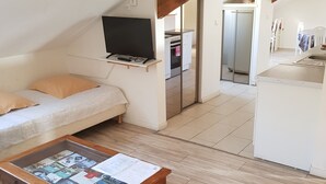 A cozy, well-lit room with a wooden floor, a sofa with cushions, a coffee table with magazines, a TV on a shelf, and a kitchen area with a refrigerator and stove.