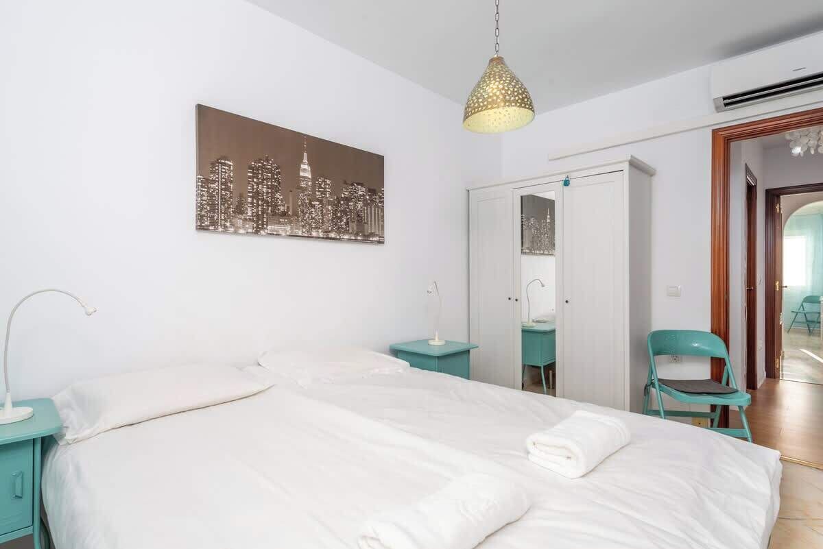 Ein Schlafzimmer mit einem Bett, einem Stadtbildgemälde, einer Pendelleuchte, einem Spiegel und einer Tür.