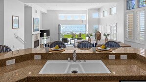 A modern kitchen with granite countertops, a white sink, and wooden cabinets. There is a dining area with a table and chairs, and a view of the sea through large windows. - LOWER LEVEL Pet Friendly Oceanfront Condo P919-0 (Oceanside)