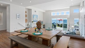 A spacious dining area with a wooden table and benches, a view of the ocean through large windows, and a balcony with outdoor seating. - LOWER LEVEL Pet Friendly Oceanfront Condo P919-0 (Oceanside)