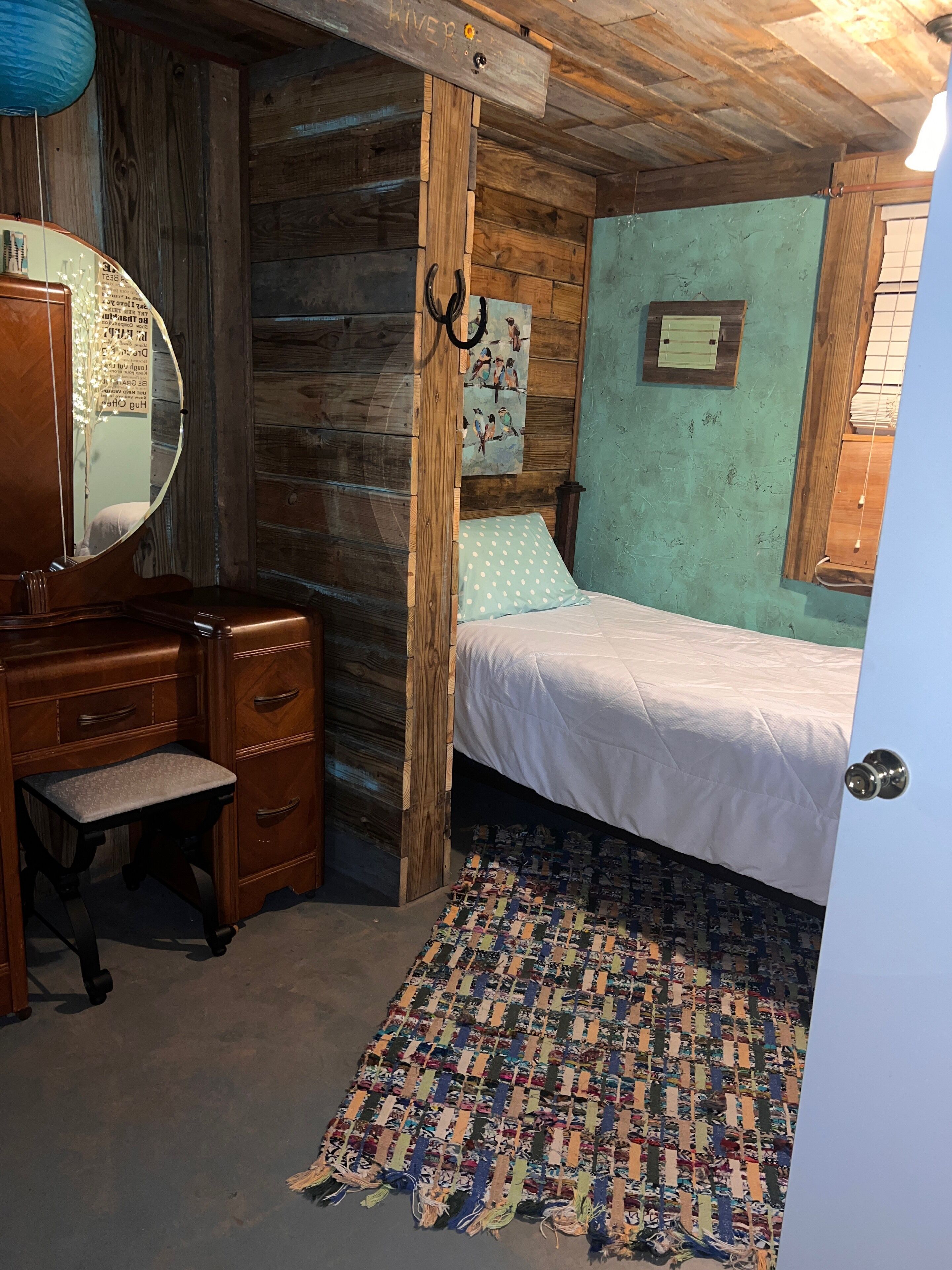 Een slaapkamer met een houten hoofdbord, een bed met een wit laken, een houten dressoir met lades, een spiegel en een aan de muur hangend schilderij.