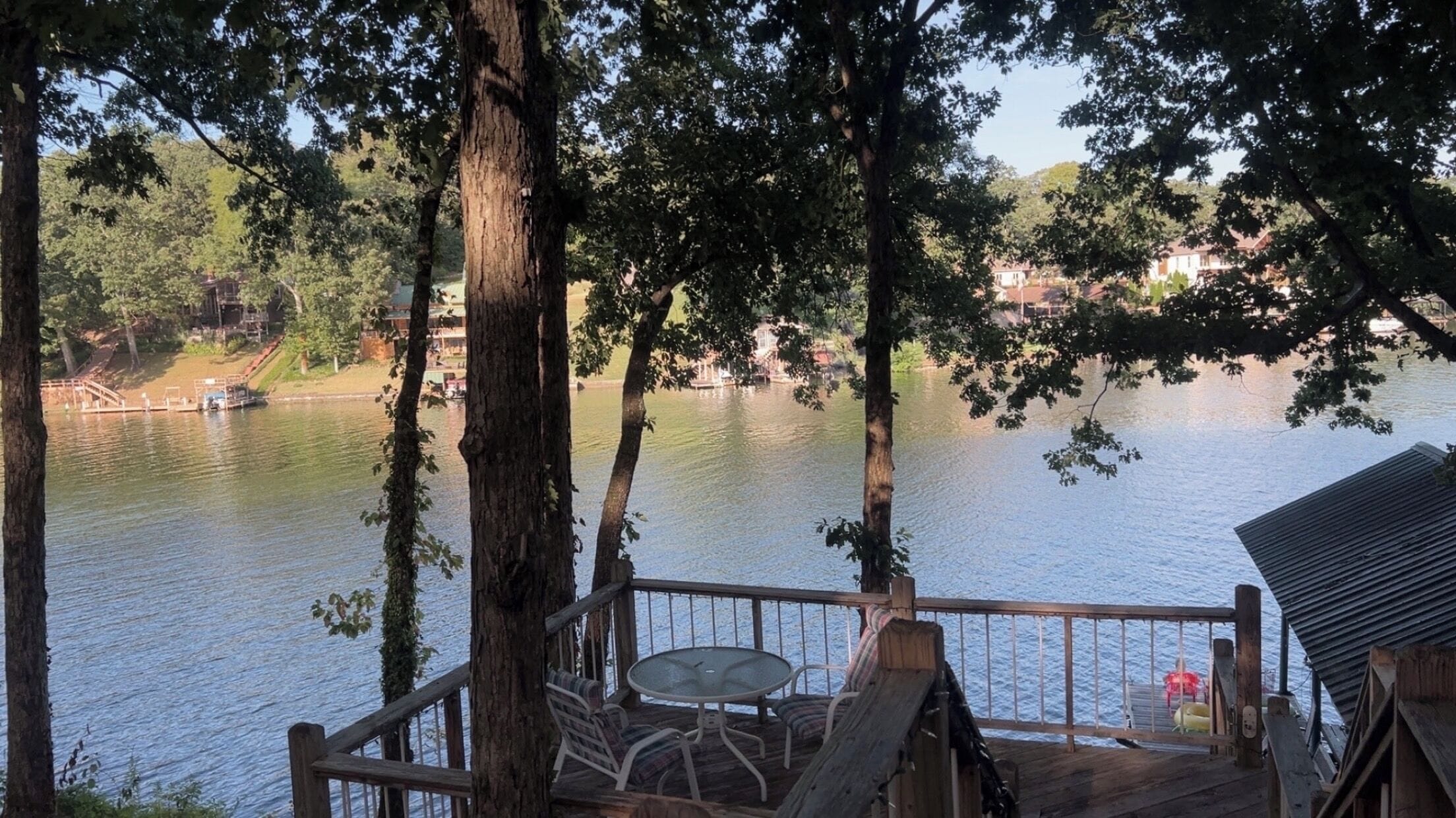 A wooden deck with a table and chairs overlooking a calm lake, surrounded by trees.