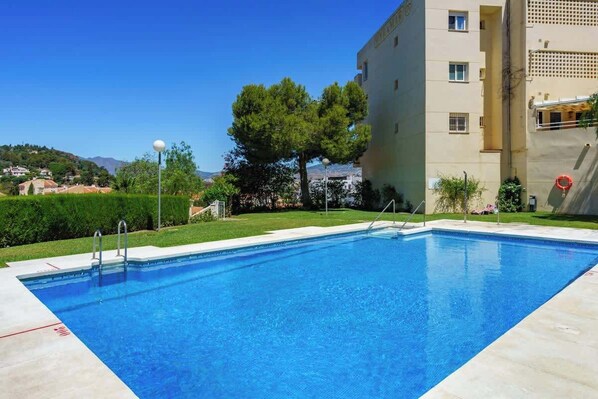 A rectangular swimming pool with clear blue water, surrounded by a paved deck. There is a building with a beige facade and a balcony on the right side.