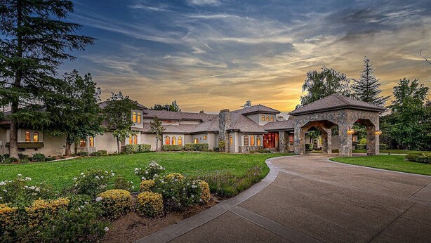 A residential property with a curved driveway, a stone archway, and a well-maintained lawn with various plants.