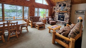Interior acogedor de una cabaña con chimenea de piedra, muebles de madera y grandes ventanales con vistas a un bosque.