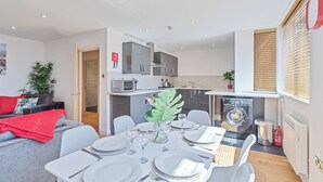 Una cocina moderna con horno empotrado, microondas y lavadora. Hay un comedor con una mesa dispuesta para cuatro personas y una sala de estar con sofá y una manta roja.