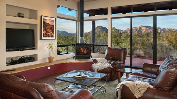 A spacious living room with leather furniture, a fireplace, a flat-screen TV, and a large window offering a mountain view. - Walls of Glass - Panoramic Views, Hot Tub, Fire Pit Under the Stars (Sedona)