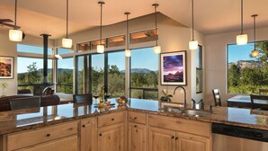 A modern kitchen with granite countertops, wooden cabinets, and a large island. There are pendant lights, a built-in stove, and a view of the outdoors through large windows. - Walls of Glass - Panoramic Views, Hot Tub, Fire Pit Under the Stars (Sedona)