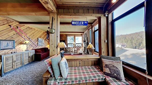 Cozy cabin interior with a rustic aesthetic, featuring a wooden ceiling, large window, and plaid-patterned seating area.