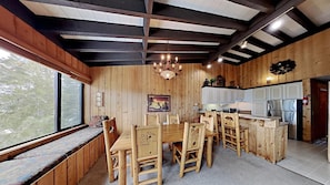 A wooden interior with a dining table and chairs, a chandelier, a kitchen area, and a window with a view of trees.