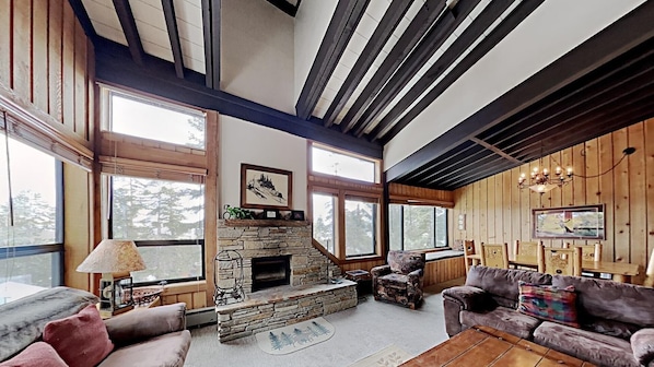 A spacious living room with a stone fireplace, large windows, wooden beams, and a cozy seating area.