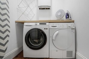 Un área de lavandería con una lavadora Samsung y una secadora Bosch, un estante de madera encima y un ventilador fijado a la pared.