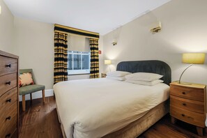 Habitación de hotel con cama, mesita de noche, lámpara, silla, cómoda y ventana con cortinas.