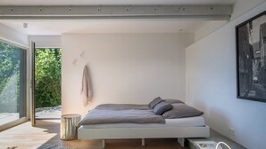 A modern bedroom with a minimalist design, featuring a bed with gray bedding, a small bedside table, and a large window providing a view of greenery outside. - Modern Minimalistic Seeblick Design Studio mit Küche, Terrace und Parkplatz (Sipplingen)