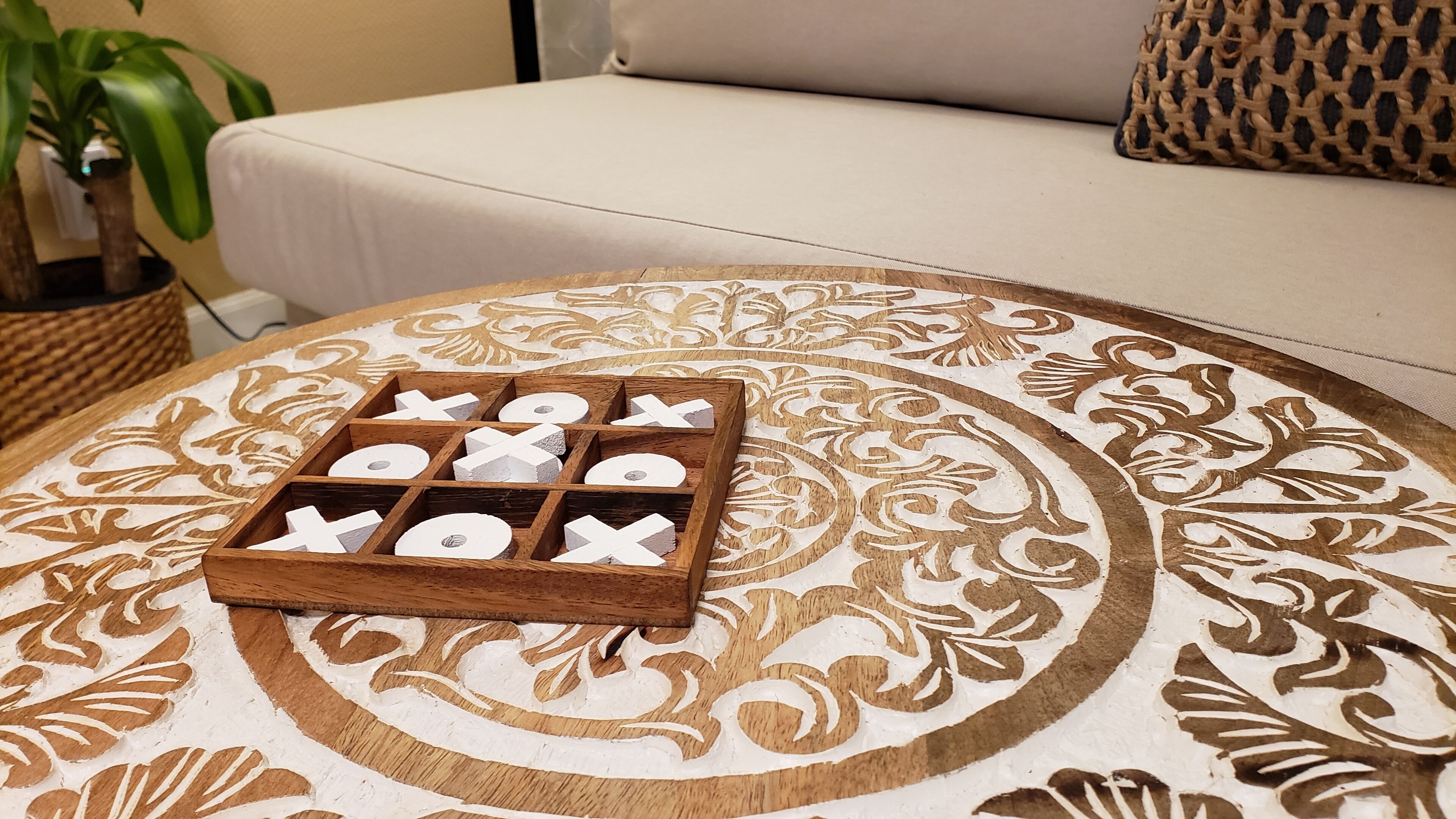 Una bandeja de madera con letras recortadas sobre un mantel estampado, colocada sobre una mesa redonda en una habitación con un sofá color beige y una cesta tejida.