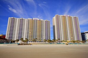 Dos hoteles de gran altura con varios balcones, una playa en primer plano y un cielo azul despejado.