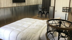 Huoneessa on sänky, seinälle asennettu televisio, pieni pöytä tuoleineen ja ovi.
