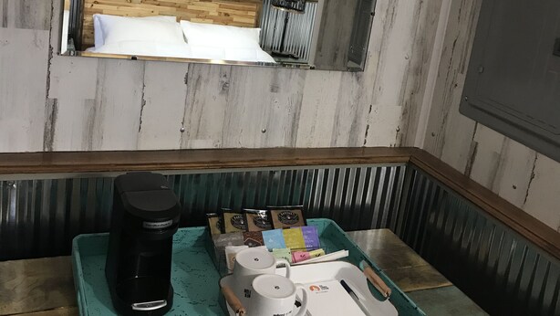 Huoneessa on puinen sängynpääty, peili, kahvinkeitin, tarjotinalusta, jossa on kahvipannu, kuppeja ja laatikko paperiliinoja.