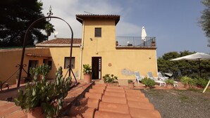 A two-story building with a terracotta facade, a balcony with a white umbrella, and a paved entrance with steps. - Lemon's house by the sea (Sicilia)