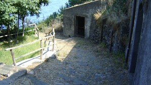 A stone pathway leading to a rustic building with a barred door, surrounded by greenery and a body of water in the background. - Lemon's house by the sea (Sicilia)