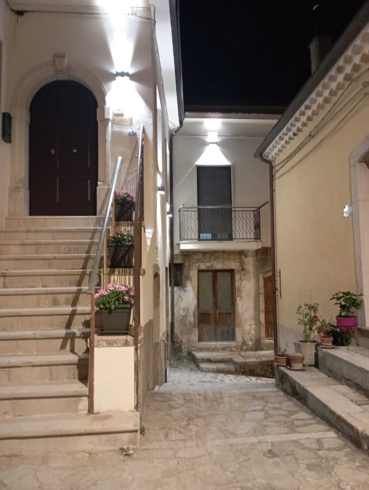 Ein schmaler Gang mit einer Treppe zu einer Tür, einem Balkon mit Geländer und Topfpflanzen.