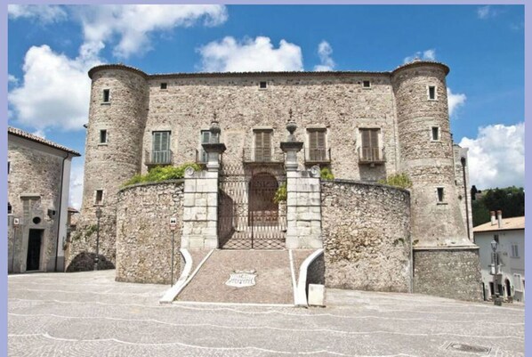 A historic stone building with a central entrance, flanked by two towers, and a paved courtyard.