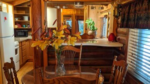 A wooden cabin interior with a dining table, chairs, a refrigerator, a microwave, and a kitchen area. - Cozy Log Cabin in peaceful wooded surroundings (Monroe)