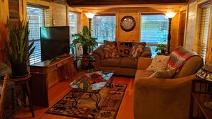 A cozy living room with a wooden interior, a television, a sofa, a coffee table, and a large window with blinds. - Cozy Log Cabin in peaceful wooded surroundings (Monroe)