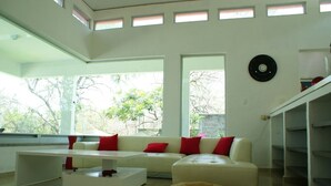 Un salon moderne avec un canapé blanc, des coussins rouges, une table basse blanche, un pouf beige et une grande fenêtre donnant sur la forêt.