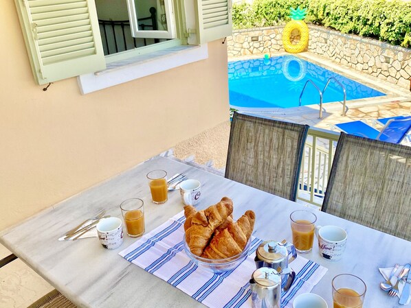 A table set with croissants, coffee cups, and a teapot, overlooking a swimming pool with lounge chairs.