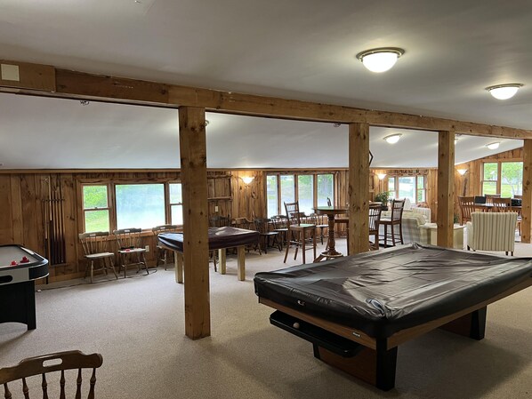 A spacious indoor area with a pool table, multiple tables and chairs, and large windows providing natural light.