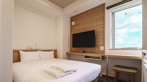 A neatly made bed with pillows and folded towels, a flat-screen TV mounted on a wooden panel above a desk, and a window with a view of the sky.