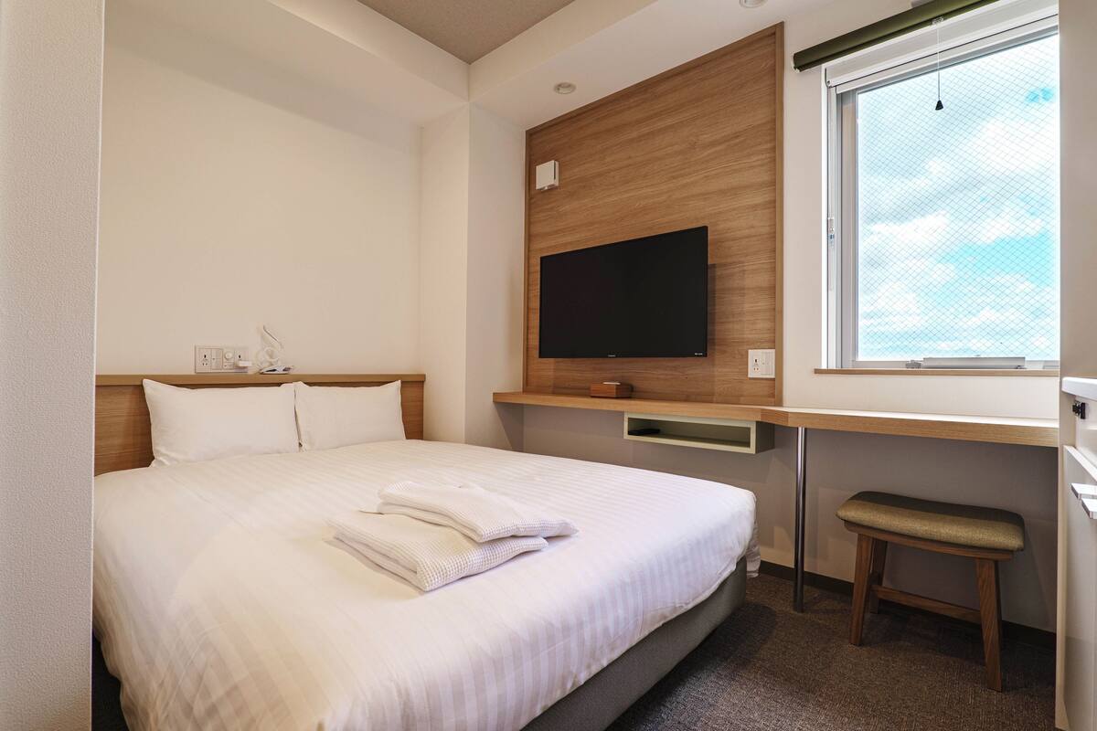 Una cama bien hecha con almohadas y toallas dobladas, un televisor de pantalla plana montado sobre un panel de madera sobre un escritorio, y una ventana con vista al cielo.