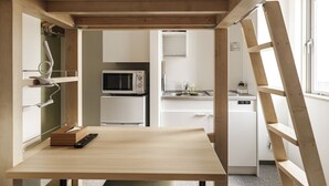 A compact kitchen with a microwave, sink, and cabinets. A wooden table with a remote control is in the foreground.
