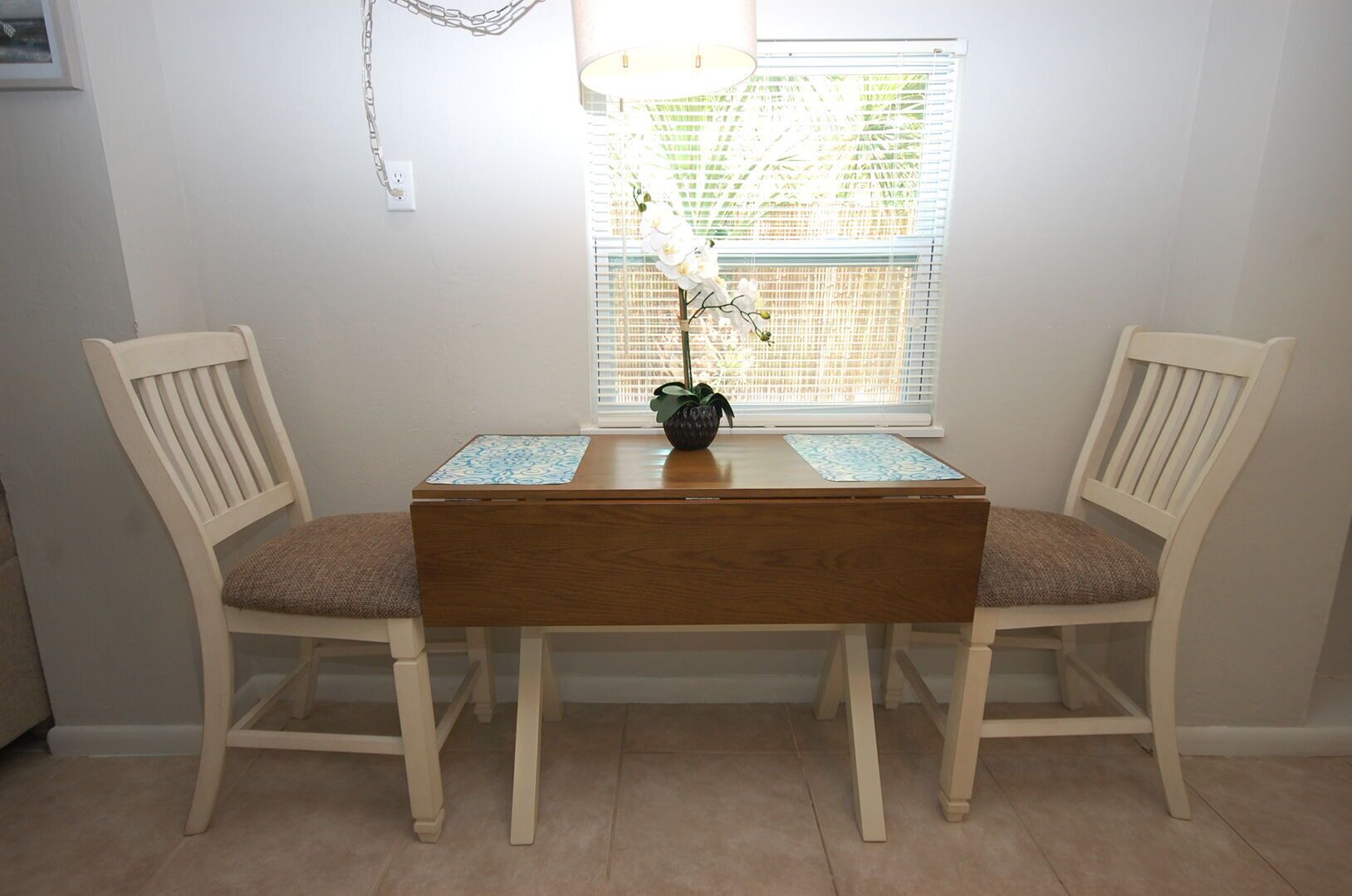 Une table à manger avec deux chaises, un vase rempli de fleurs et une fenêtre avec des stores.