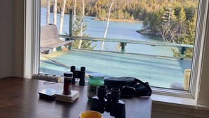 A table with binoculars, a mug, and a croissant, overlooking a lake and forest.