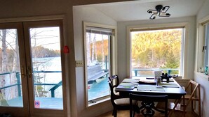 A room with a wooden table, chairs, and large windows offering views of a dock and trees.