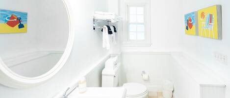 A bathroom with a large round mirror, a white sink, a toilet, and a small window. There are two paintings on the wall.