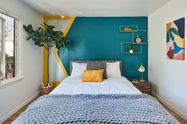 Quarto com cama, mesa de cabeceira, abajur, planta e janela.