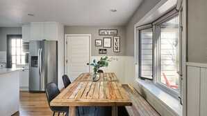 Une cuisine avec une table en bois, des chaises, un réfrigérateur et une fenêtre avec des stores.