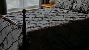 Ein Schlafzimmer mit einem Bett, einem Nachttisch, einer Lampe und einem Fenster mit Vorhängen.