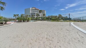 Ein Strandabschnitt mit Sand, im Hintergrund ein großes Gebäude, Palmen und ein Volleyballnetz.