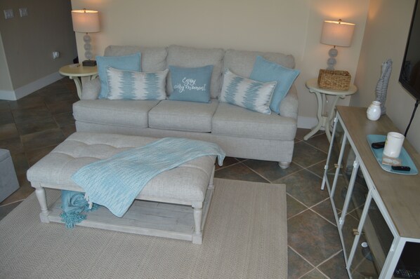 Un salon avec un canapé gris, un pouf assorti et une petite table d’appoint sur laquelle se trouve une lampe.