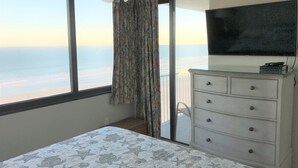 Een slaapkamer met een groot raam, een plafondventilator, een bed met een beddekleed met dessin, een dressoir en een aan de muur bevestigde televisie.