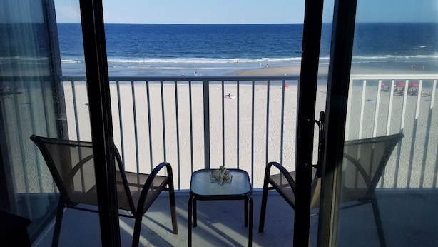 Een balkon met twee stoelen en een kleine tafel, met uitzicht op een strand met mensen en parasols.