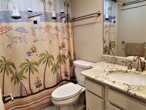 A bathroom with a beach-themed shower curtain, a toilet, a sink with a marble countertop, and a towel rack.