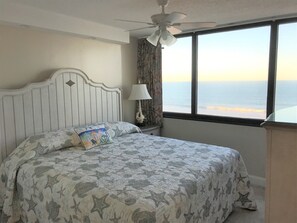 A bedroom with a bed, a ceiling fan, a window with a view of the ocean, and a lamp.