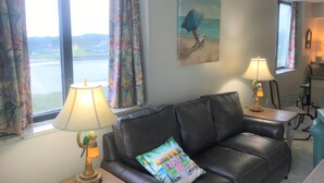 Een woonkamer met een zwarte leren bank, twee lampen, een kleurrijk kussen, een schilderij van een zeilboot en uitzicht op een wateroppervlak door grote ramen.