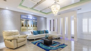 A spacious living room with a beige sofa, a recliner, and a coffee table. There is a large abstract painting on the wall and a chandelier hanging from the ceiling.