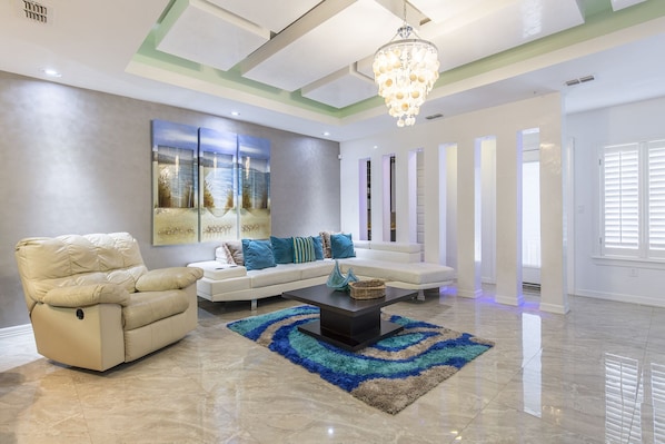 Un salon spacieux avec un canapé beige, un fauteuil inclinable et une table basse. Un grand tableau abstrait orne le mur, et un lustre est suspendu au plafond.